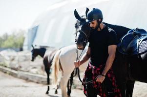 El hombre árabe de barba alta usa casco negro con caballo árabe. foto