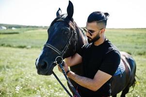 hombre árabe de barba alta vestido de negro y gafas de sol con caballo árabe. foto