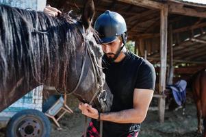 El hombre árabe de barba alta usa casco negro con caballo árabe. foto