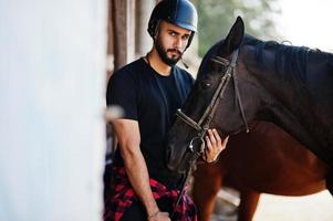 El hombre árabe de barba alta usa casco negro con caballo árabe. foto