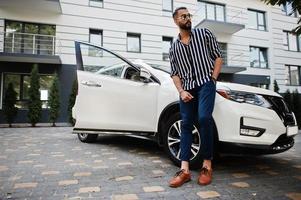 Successful arab man wear in striped shirt and sunglasses pose near his white suv car. Stylish arabian men in transport. photo