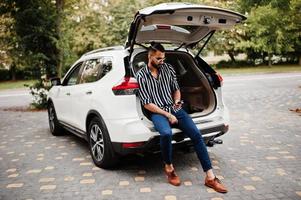 Successful arab man wear in striped shirt and sunglasses sitting in the trunk of his white suv car with mobile phone. photo