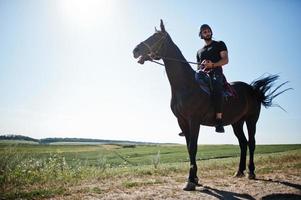 El hombre árabe de barba alta usa casco negro, monta un caballo árabe. foto