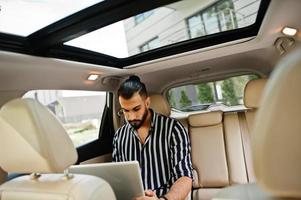 Successful arab man wear in striped shirt and sunglasses pose inside white suv car with laptop in hands. photo