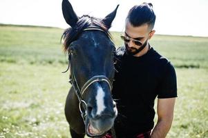 Arab tall beard man wear in black and sunglasses with arabian horse. photo