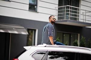 Successful arab man wear in striped shirt and sunglasses sitting on the roof of his white suv car. photo