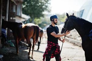 El hombre árabe de barba alta usa casco negro con caballo árabe. foto
