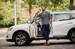 Successful arab man wear in striped shirt and sunglasses pose near his white suv car. Stylish arabian men in transport. photo