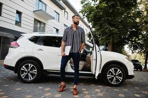 Successful arab man wear in striped shirt and sunglasses pose near his white suv car with laptop in hands. photo