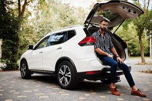 Successful arab man wear in striped shirt and sunglasses sitting in the trunk of his white suv car. Stylish arabian men in transport. photo