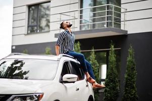 Successful arab man wear in striped shirt and sunglasses sitting on the roof of his white suv car. photo