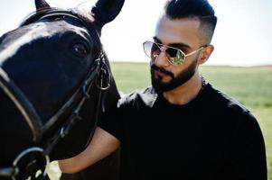 Arab tall beard man wear in black and sunglasses with arabian horse. photo