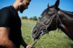 Hombre árabe de barba alta vestido de negro con caballo árabe. foto