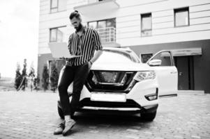 Successful arab man wear in striped shirt and sunglasses pose near his white suv car with laptop in hand. photo