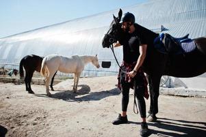 El hombre árabe de barba alta usa casco negro con caballo árabe. foto