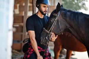 El hombre árabe de barba alta usa casco negro con caballo árabe. foto