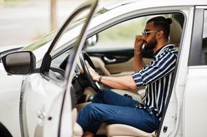 Successful arab man wear in striped shirt and sunglasses pose behind the wheel of  his white suv car. Stylish arabian men in transport. photo