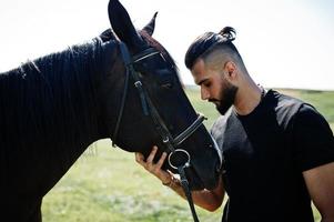 Hombre árabe de barba alta vestido de negro con caballo árabe. foto