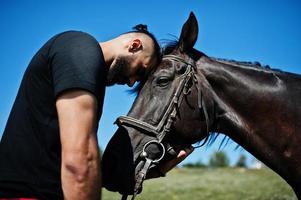 Hombre árabe de barba alta vestido de negro con caballo árabe. foto