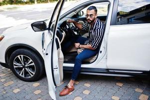 Successful arab man wear in striped shirt and sunglasses pose behind the wheel of  his white suv car. Stylish arabian men in transport. photo