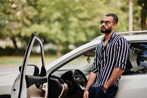 Successful arab man wear in striped shirt and sunglasses pose near his white suv car. Stylish arabian men in transport. photo