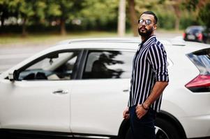 Un exitoso hombre árabe vestido con camisa a rayas y gafas de sol posan cerca de su auto todoterreno blanco. hombres árabes con estilo en el transporte. foto