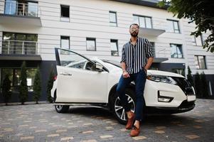 Successful arab man wear in striped shirt and sunglasses pose near his white suv car. Stylish arabian men in transport. photo