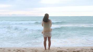 retrato de uma linda mulher asiática sorrindo relaxante na praia. uma jovem passeando à beira-mar olhando as grandes ondas e ventos fortes. conceito de relaxamento e viagens de férias. video