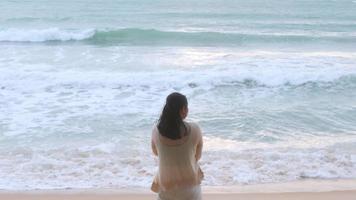 portrait d'une belle femme asiatique souriante relaxante sur la plage. une jeune femme se promenant le long de la mer en regardant les grosses vagues et les vents forts. concept de détente et de voyage en vacances. video