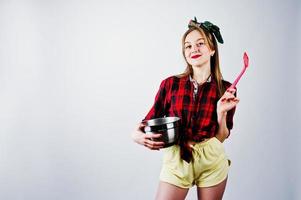 Young funny housewife in checkered shirt and yellow shorts pin up style with saucepan and kitchen spoon isolated on white background. photo