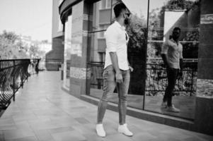 Stylish tall arabian man model in white shirt, jeans and sunglasses posed at street of city. Beard attractive arab guy against modern building. photo