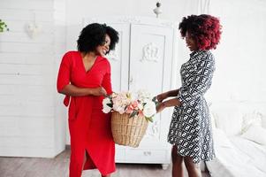 Two fashionable african american woman in evening dress standing with flowers basket on hands against old vintage wardrobe at white room. photo