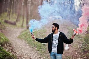 hombre árabe de estilo callejero con anteojos sostiene bengalas de mano con bomba de granada de humo rojo y azul. foto