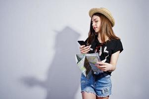 chica turista con mapa, vestida con camisa jajaja, pantalones cortos y sombrero hablando por teléfono móvil aislado en blanco. foto