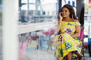 linda chica afroamericana de pequeña altura con rastas, vestida con un vestido amarillo de color, sentada en un café en el centro comercial y hablando por teléfono. foto