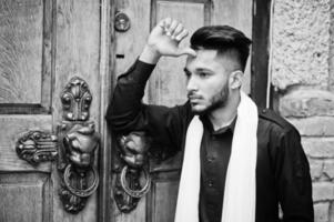 Indian stylish man in black traditional clothes with white scarf posed outdoor against old gates. photo