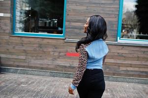 Hipster african american girl wearing jeans shirt with leopard sleeves posing at street against wooden house with windows. photo