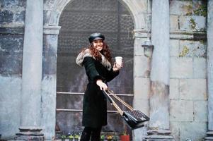 chica mexicana rizada con gorra de cuero y taza de café de plástico a mano caminando por las calles de la ciudad. foto