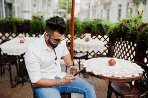 Stylish tall arabian man model in white shirt, jeans and sunglasses posed at street of city. Beard attractive arab guy sitting on outdoor cafe and looking at mobile phone. photo
