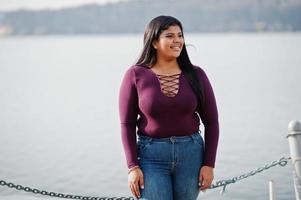 Pretty latino xxl model girl from Ecuador wear on violet blouse posed against lake. photo