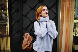 Cheerful young beautiful redhaired woman in glasses, warm blue wool sweater with backpack posed outdoor. photo