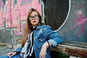 chica hipster casual con estilo en ropa de jeans y gafas contra una gran pared de graffiti. foto