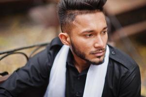 Indian stylish man in black traditional clothes with white scarf posed outdoor sitting on bench. photo