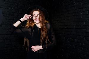 sesión de estudio de una chica de negro con rastas, anteojos y sombrero sobre fondo de ladrillo. foto