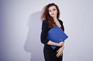 Brunette curly haired girl in black office jacket with skirt, on bra with folder of documentts at hands on studio against white background. Sexy businesswoman. photo