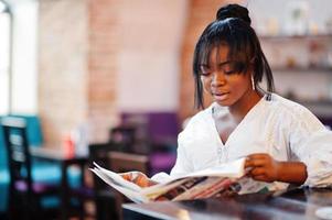 Stylish african american women in white blouse and blue jeans posed at cafe with newspaper. photo