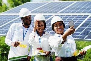 African american technician checks the maintenance of the solar panels. Group of three black engineers meeting at solar station. Make selfie by phone. photo