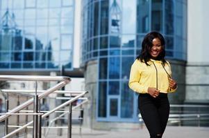 Hipster african american girl wearing yellow hoodie posing at street against office building with blue windows. photo