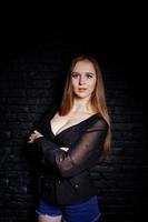 Studio shot of brunette girl in black blouse with bra and shorts against black brick wall. photo