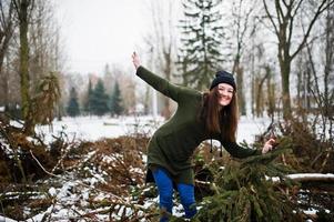 Young girl wear on long green sweatshirt, jeans and black headwear at  branches of the pine tree in winter day. photo